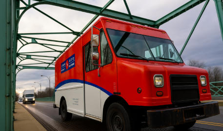 A red and white Canada Post delivery truck crossing a green steel bridge under cloudy skies.
